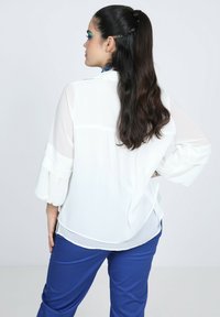 Weiße, durchsichtige Bluse mit langen, leicht ausgestellten Ärmeln und fließendem Design, getragen mit blauen Hosen, die einen gestuften hinteren Saum haben.