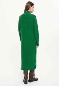 Langes grünes Strickkleid mit hohem Kragen, Rippstruktur, seitlichen Schlitzen und langen Ärmeln. Getragen mit braunen Stiefeln bis zum Knie.