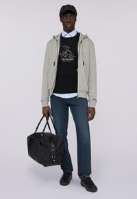 Sudadera gris con cremallera sobre un suéter negro con logo, cuello de camisa azul, jeans oscuros, zapatos negros y una gran bolsa de viaje negra.