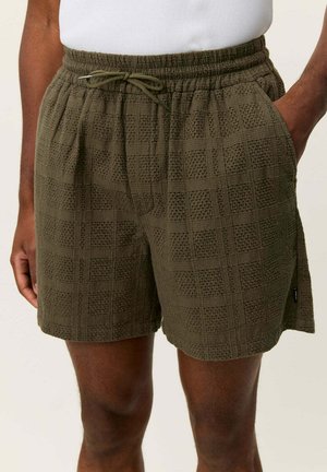 Mørke olivengrønne teksturerte shorts med snor i midjen, båret av en person med hendene i lommene mot en ensfarget bakgrunn.