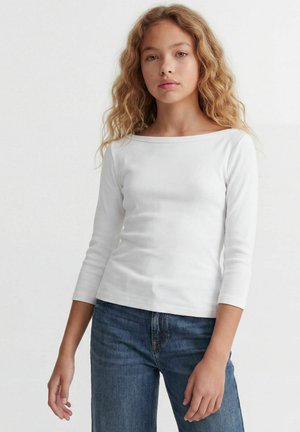 Une femme porte un haut blanc ajusté à manches longues et un jean bleu sur un fond clair uni.
