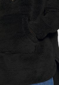 Sort fuzzy jakke med hånd gemt i sidelommen, båret over blå denimjeans.