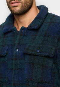 Giacca in pile con bottoni in un motivo a quadri verde scuro e blu. Dotata di due tasche frontali e colletto, realizzata in un tessuto morbido e testurizzato.