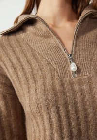 Gros plan sur une personne portant un pull en maille côtelée marron avec une fermeture éclair argentée munie d'une grande tirette en perle blanche.