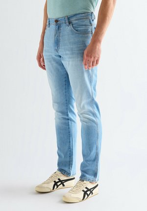 Mann trägt hellblaue, schlanke Jeans und cremefarbene Sneakers mit schwarzen Streifen, steht vor einem weißen Hintergrund.