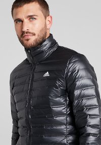 Czarna kurtka puchowa z pionowym pikowaniem, wysoki kołnierz i zamek błyskawiczny. Posiada małe białe logo Adidas na górze po lewej stronie.