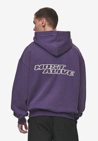 Sudadera morada de algodón con un corte relajado. Presenta un gran gráfico blanco que dice "MOST ALIVE" impreso en la parte trasera. La parte delantera es lisa.