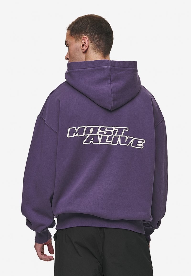 Sudadera morada de algodón con un corte relajado. Presenta un gran gráfico blanco que dice "MOST ALIVE" impreso en la parte trasera. La parte delantera es lisa.
