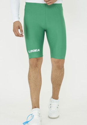 Uomo con pantaloncini sportivi verdi fino al ginocchio con logo LEGEA e scarpe da ginnastica bianche, in piedi davanti a uno sfondo uniforme.