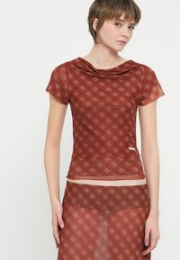 Sheer, short-sleeved top in a rust red with a plaid pattern and draped neckline. Features an underlayer and subtle hardware accent.