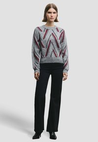 Naine, kes kannab halli kampsunit punaste ja mustade siksakmustritega, musti laia säärega pükse ning musti kontsaga pahkluu-saapaid, seisab neutraalse tausta ees.