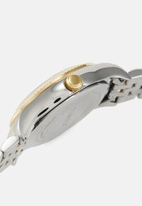 Vue latérale d'une montre-bracelet en métal bicolore montrant une couronne dorée, un boîtier argenté, et un bracelet en métal argenté et doré, sur fond blanc.