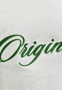 Zöld texturált kézírás "Origin" felirattal, világosszürke textil háttéren hímzett.