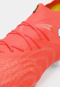 Lyse rød fodboldstøvle med struktureret overdel, matchende røde skosnørebånd og et lille sort og gult logo nær anklen.