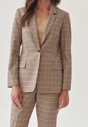 Kobieta ubrana w beżowo-brązową kratowaną marynarkę i dopasowane spodnie oraz jasnobrązowy top pod spodem, stojąca na jednolitym tle.