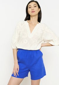 Balta izšūta blūze ar V-veida kakla izgriezumu un piepūšamām piedurknēm, kombinēta ar ziliem linu šortiem. Iespējami ziedu raksti un elastīga jostu.