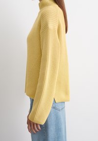Żółty sweter dzianinowy z wysokim kołnierzem, wyróżniający się teksturowanym wzorem i opuszczonymi ramionami. Skrócony krój z bocznymi rozcięciami dla komfortu.