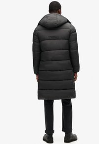 Długa czarna puchowa kurtka z kapturem, z pikowanym wzorem i widocznym logo na plecach. Noszona z ciemnymi spodniami i butami.