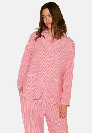 Kvinde iført løstsiddende pyjamassæt med lodrette pink og hvide striber, med knappet bluse og lommer, stående mod en ensfarvet baggrund.