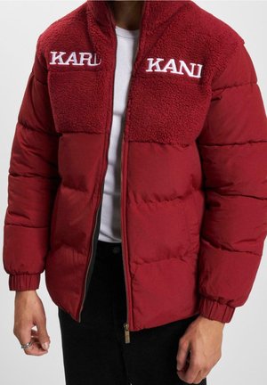Czerwona kurtka puchowa z kołnierzem z polaru i białym napisem „KARL KANI”, noszona na białej prążkowanej koszulce i czarnych spodniach.