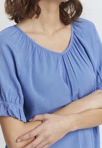 Niebieska bluzka z krótkimi bufkami, dekoltem w serek i marszczeniami na ramionach. Gładka tekstura i lekki materiał.