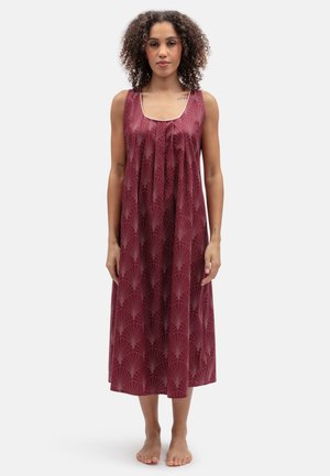 Frau mit lockigem Haar, steht barfuß, trägt ein ärmelloses bordeauxfarbenes Kleid mit leichtem geometrischem Muster vor weißem Hintergrund.