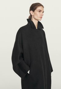 Kvinde med lys hud og brunt hår trukket tilbage, iført en sort, oversize frakke med vide ærmer og høj krave, hænderne i lommerne, vendt mod højre.