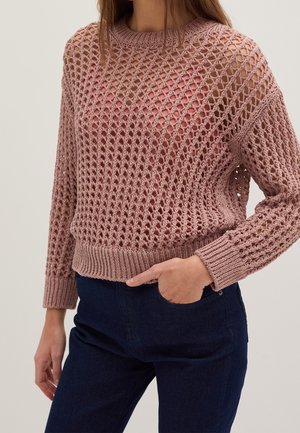 Nainen, joka pukeutunut vaaleanpunaiseen, avoimeen neulepuseroon, jossa on pitkät hihat, ruusunsävyisen topin päällä, yhdistetty tummansinisiin farkkuihin, käsi taskussa.