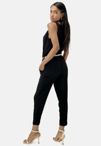Mono sin mangas negro con un corte holgado, cintura entallada que destaca un cinturón metálico trenzado, combinado con unas stilettos beige.