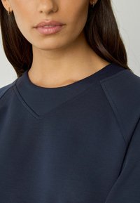 Frau, die einen marineblauen Sweatshirt mit V-förmiger gesteppter Detail am runden Halsausschnitt und kleinen goldenen Creolen trägt.