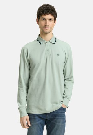 Man draagt een lichtgroen poloshirt met lange mouwen en een gestreepte kraag, gecombineerd met een blauwe spijkerbroek. Hij staat met één hand in zijn broekzak.