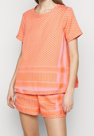 Mujer con conjunto a juego de top y pantalones cortos de manga corta naranjas con diseños geométricos, posando frente a un fondo liso y claro.