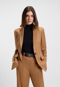 Blazer v barvě velbloudí s přizpůsobeným střihem, nošený přes černý žebrovaný rolák. Obsahuje šikmé kapsy a černý pásek s kovovými doplňky.