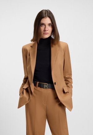 Blazer color cammello con vestibilità tailored, indossato sopra un dolcevita nero a coste. Presenta tasche inclinate e una cintura nera con accessori metallici.