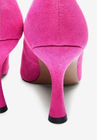 Une paire de chaussures à talons hauts en suede rose vif vue de l'arrière, mettant en valeur les talons fins et les embouts de talon noirs.