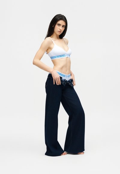 Γυναίκα που προσαρμόζει ανοιχτό φαρδύ σκέλος σκούρων τζιν, φοράει λευκό μπραλέτα και εσώρουχο με μπλε μοτίβο ελληνικού κλειδιού στις ταινίες, στέκεται πάνω σε λευκό φόντο.