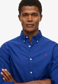 Blauwe overhemdd met een standaardkraag, witte knopen en een klein rood logo op de linkerborst. Gladde katoenen textuur.