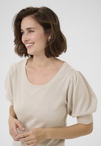 Beige svetr s texturou a výstřihem do V, se volumózními rukávy. Tkanina má mírný třpyt, což dodává designu hloubku.