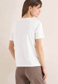 Femme aux cheveux bruns raides, vêtue d'un t-shirt blanc à manches courtes uni et d'un pantalon marron, légèrement tournée de côté sur un fond clair.