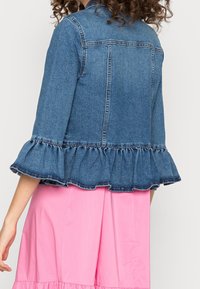 Giacca di jeans con orlo arricciato, caratterizzata da un lavaggio blu, maniche a tre quarti e colletto classico. Indossata sopra un indumento rosa.