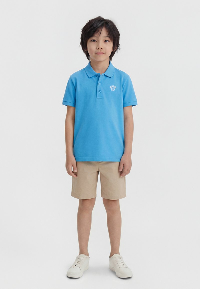 Αγόρι που στέκεται, φορώντας μπλε πόλο με λογότυπο, μπεζ σορτς και λευκά αθλητικά παπούτσια, μπροστά σε απλό φωτεινό φόντο.