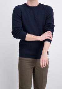 Marinblå texturerad tröja med rund hals och ribbade muddar, kombinerad med ljusbruna byxor som visar ett geometriskt mönster.