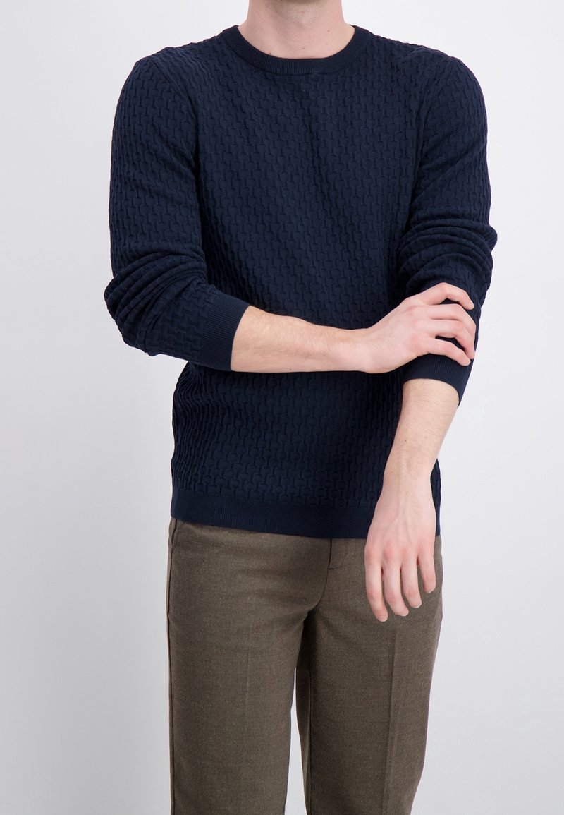 Marinblå texturerad tröja med rund hals och ribbade muddar, kombinerad med ljusbruna byxor som visar ett geometriskt mönster.