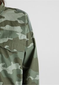 Manga y parte superior de pecho de chaqueta de camuflaje verde y marrón con bolsillo con solapa, costura visible y textura de la tela sobre un fondo liso.
