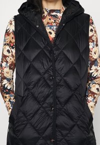 Černá vyp padded vesta s vzorem diamantového prošívání, s kapucí a předními knoflíky. Nosená přes květinovou blůzu s dlouhým rukávem v různých barvách.