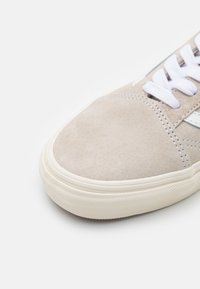 Beige suede sneaker with white laces, rounded toe, and textured rubber sole. Features subtle stitching and a smooth accent stripe.