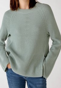 Femme portant un pull en tricot vert clair avec des zippers sur les côtés et un jean bleu, les mains dans les poches, coupé au niveau du cou et des hanches.