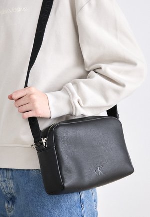 Personne portant un sweat-shirt Calvin Klein beige clair et un jean bleu, tenant un sac bandoulière en cuir noir Calvin Klein avec logo et sangle réglable.