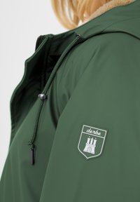 Giacca impermeabile verde con colletto foderato di pelliccia, dotata di una toppa con logo sulla manica, cordini regolabili e superficie liscia.