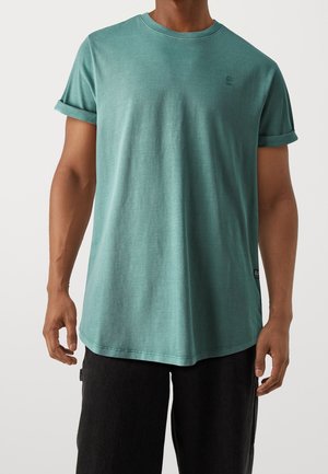 Mand iført en teal crew-neck kortærmet T-shirt med opsmøgede ærmer og sorte bukser, stående mod en ensfarvet baggrund.